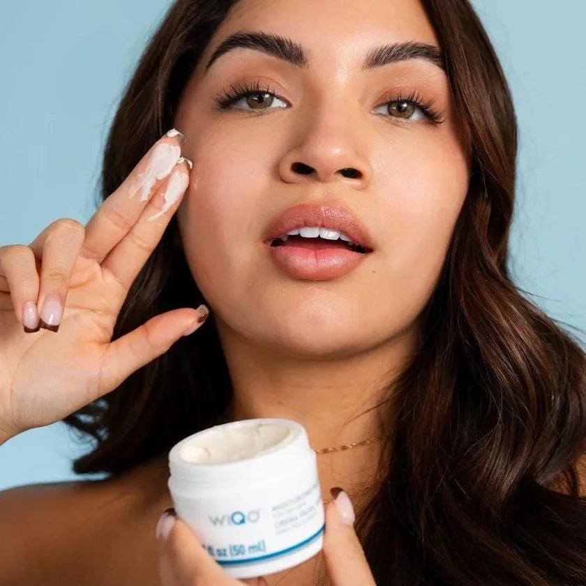 Close-up of a woman applying WiQo Moisturizing Face Cream For Dry Skin near her cheek, holding the open jar below.